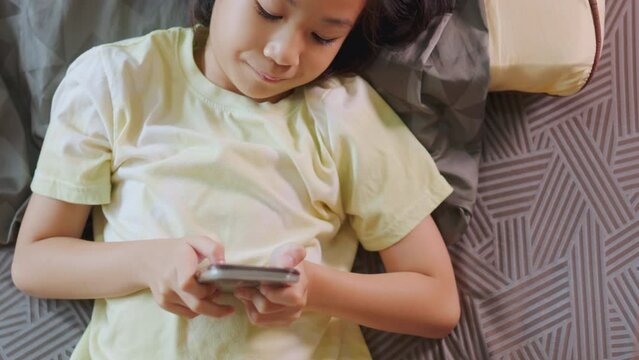 Top view of adorable Asian kid girl, lied in bed, enjoys her smartphone in a sunlit room. Her expression hints at digital fun, highlighting the allure of home tech, playing game and e-learning.