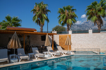 a luxurious pool area with loungers and umbrellas, a clear blue pool reflecting the sky, and a mountainous backdrop. Palm trees add to the resort-like ambiance