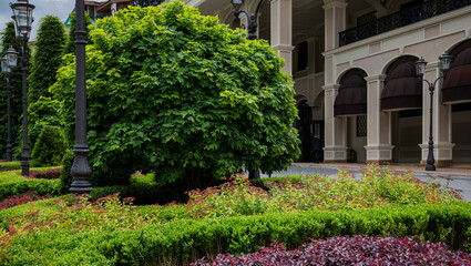 a landscaped pathway in an upscale outdoor area, a shopping or hotel complex. The path is flanked by lush greenery and ornamental shrubs. Street lamps line the walkway, adding to the elegant ambiance