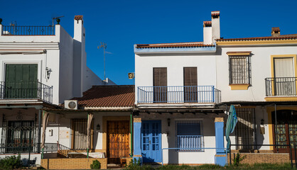a row of charming traditional Andalusian townhouses, with white walls and terracotta roofs, featuring colorful doors and wrought-iron balconies under a clear, vivid blue sky