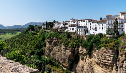 Fototapeta premium Perched atop a dramatic cliff, the white buildings of a Mediterranean town bask in the sunlight, overlooking lush green valleys and distant rolling hills under a clear blue sky