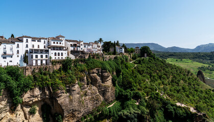 Fototapeta premium Perched atop a dramatic cliff, the white buildings of a Mediterranean town bask in the sunlight, overlooking lush green valleys and distant rolling hills under a clear blue sky