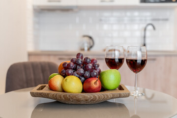fruits on a wooden plate and two glasses of red wine on the kitchen table. White modern kitchen. Ripe apples, grapes, tangerines and pears on the table.