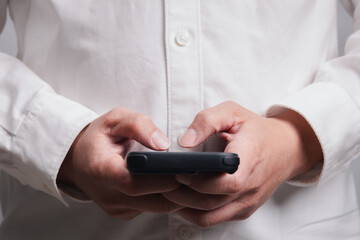 Fototapeta premium A man is holding a cell phone in his hand. He is wearing a white shirt and he is focused on his smartphone in concept of mobile technology.