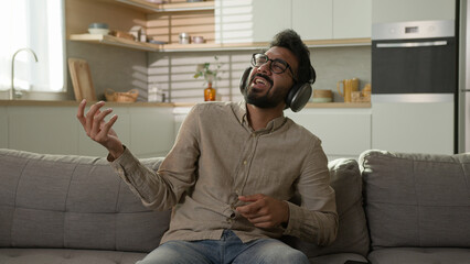 Happy relaxed Arabian man homeowner listen music in headphones enjoy rhythm song pretend playing drums on home sofa cheerful Indian guy listening musical track in earphones energetic dance in kitchen