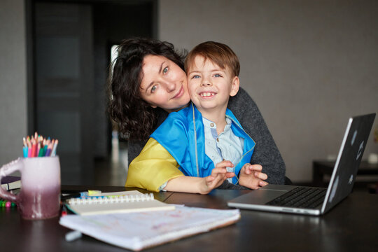 Ukrainian mother and son with flag sitting with laptop at home studying .Happy smiling family hugs and looking at the camera. Ukrainian refugees concept. High quality photo