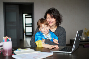 Ukrainian mother and son with flag sitting with laptop at home studying .Happy smiling family hugs and looking at the camera. Ukrainian refugees concept. High quality photo