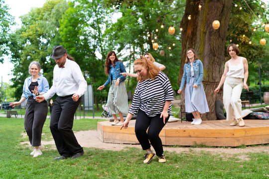 Cheerful dance party outdoors, featuring individuals of different ages enjoying the festive ambiance under illuminated string lights. They are dancing swing dance in summer city park