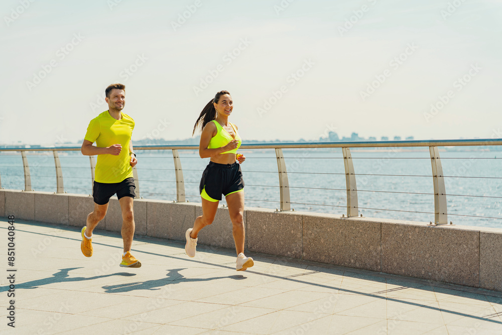 Wall mural a vibrant scene of a man and woman jogging along a seaside promenade under a clear sky.