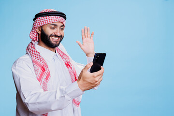 Smiling muslim man wearing traditional clothes greeting while speaking in video chat on smartphone. Arab waving hi while talking in virtual meeting on mobile phone with cheerful expression © DC Studio