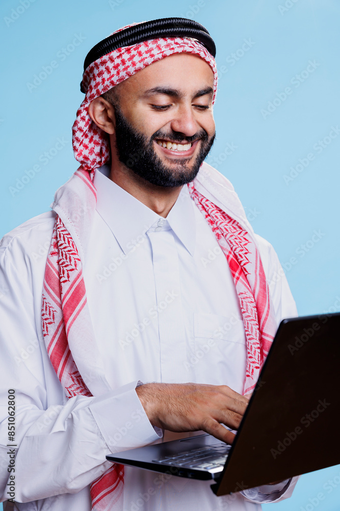 Poster smiling man wearing islamic thobe and ghutra texting in social media while standing with laptop in s
