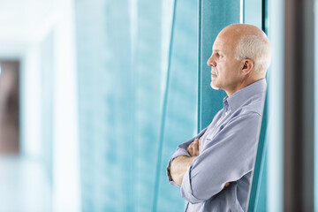 Senior businessman looking through office window with arms cross - Powered by Adobe