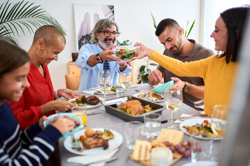 Smiling Caucasian gray-haired man sitting at table passing salad to his happy daughter at meal event indoors. Multi-generational diverse cheerful people enjoying family reunion together at weekend