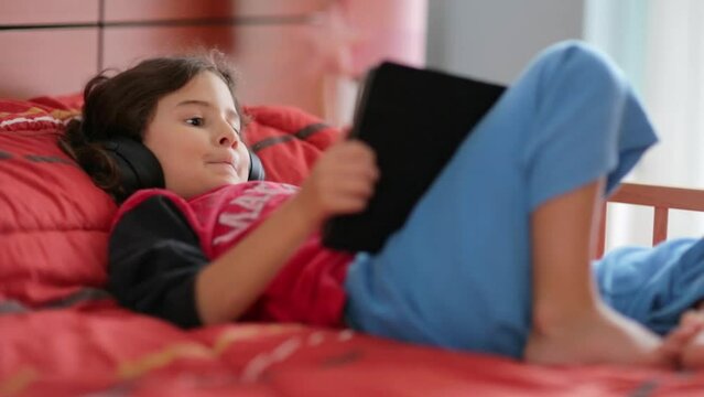 A young boy lies on a red bed, headphones on, engrossed in his digital tablet. He appears to be lost in a world of entertainment, perhaps a game or movie.