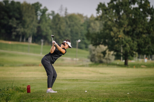 Sigulda, Latvia - September 10, 2023 - Woman golfer in black attire taking a swing on a green golf course, with trees and a red marker in the background.