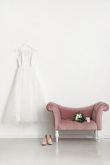 Wedding dress hanging on white wall and bridal bouquet on pink velvet armchair in room