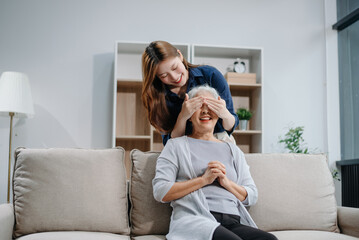 Happy adult Asian granddaughter and senior grandmother having fun enjoying talk sit on sofa in modern living room, using a laptop, tablet
