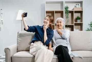 Happy adult Asian granddaughter and senior grandmother having fun enjoying talk sit on sofa in modern living room, using a laptop, tablet