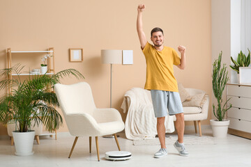 Cheerful young man with modern robot vacuum cleaner at home