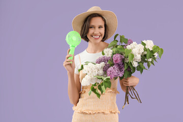 Young woman with green small electric fan and bouquet of lilac flowers on lilac background