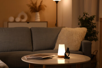Glowing lamp on coffee table, comfortable sofa and magazines in stylish living room
