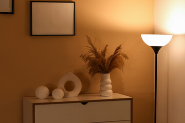 Glowing lamp near chest of drawers with vase of pampas grass in stylish living room