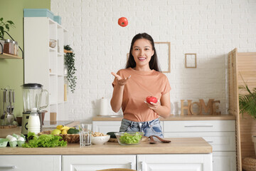 Beautiful young happy Asian woman with fresh tomatoes for salad in kitchen