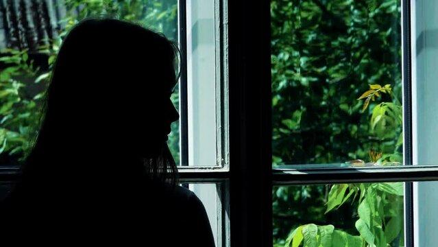 A black silhouette of a woman standing by a window, evoking feelings of loneliness and depression. The stark contrast between the dark figure and the light outside emphasizes the emotional depth