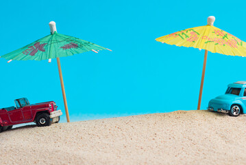 Toy car next to cocktail umbrella on sea sand, blue background. Concept of travel, vacation, summer.