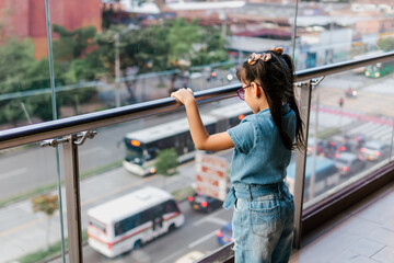 Happy and amazed Latina girl with glasses watching the cars go by on a high floor. Childhood, vacation and fun. Children day.