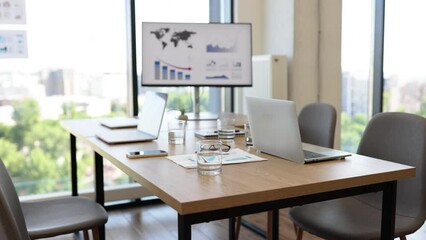 Empty office meeting room with laptops on a wooden table, a presentation screen in the background, and large windows offering a city view.