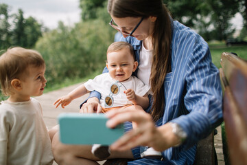 Mother entertains her kids with a mobile phone while sitting on a park bench, sharing a video call