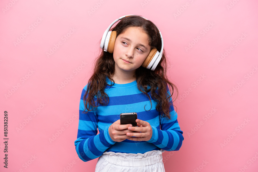 Wall mural little girl isolated on pink background listening music with a mobile and thinking