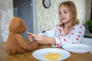 A charming blonde baby in cute pajamas with hearts is eating a delicious breakfast and feeding her...