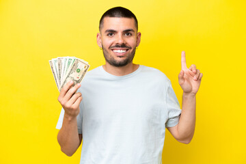 Young caucasian man taking a lot of money isolated on yellow background pointing up a great idea