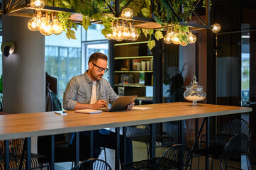 Focused male accountant in eyeglasses auditing financial reports over laptop on table in office