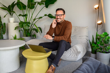 Smiling businessman discussing project details over phone call and using laptop on comfortable sofa