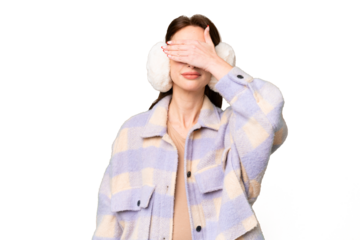 Young caucasian woman wearing winter muffs over isolated chroma key background covering eyes by hands. Do not want to see something