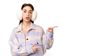 Young caucasian woman wearing winter muffs over isolated chroma key background surprised and pointing side