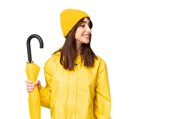 Young caucasian woman with rainproof coat and umbrella over isolated chroma key background looking to the side and smiling