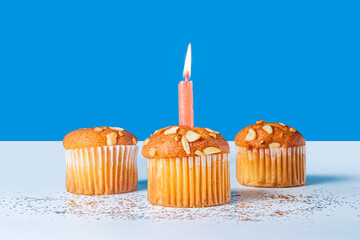 Three birthday cupcakes with single lit candle on white table on against blue background. Closeup