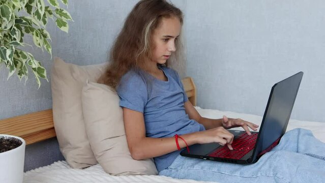 In the bedroom, on a cozy bed, a young girl is typing on her laptop, having blonde hair