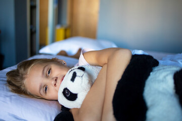 A little blonde girl with a plush toy on a huge bed in an elegantly furnished hotel room.
