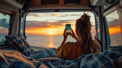 Woman Taking Photo of Sunset from Camper Van. A woman is seen inside a cozy camper van, taking a photo of a breathtaking sunset over the ocean with her phone.