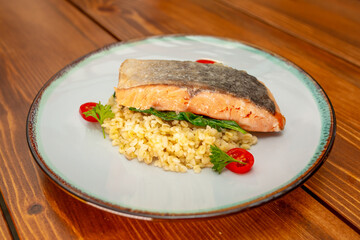 Grilled baked salmon garnished with bulgur, tomatoes, herbs and lemon. Fish dish, grilled salmon steak on a wooden table.