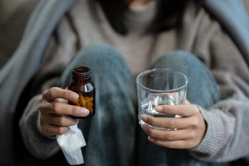 woman sitting on sofa covered with blanket freezing blowing running nose got fever caught cold, holding pills and glass of water.