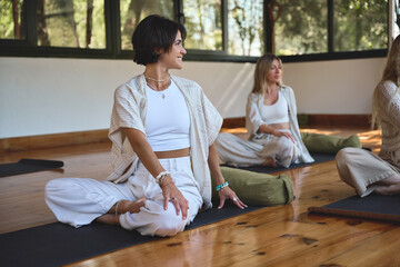 Young happy yogi women doing yoga exercises at group class yoga meditation session sitting in yoga pose on wooden floor in studio practicing body care meditation experience during retreat therapy.