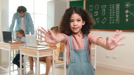 American girl looking at camera while waving hand to greeting. Group of diverse elementary student...