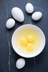 White bowl with raw chicken and goose eggs, vertical shot on a black marble background, top view