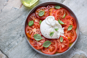 Burrata cheese with red tomato slices and green basil leaves, horizontal shot on a grey granite background, elevated view